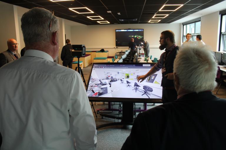 A group of people looking at a large white screen which displays a computer generated image of an office layout.